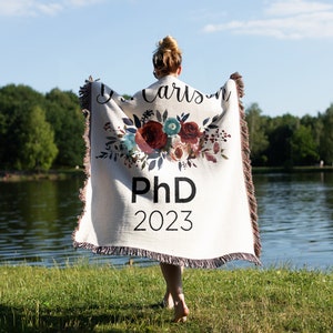 K&ouml;nnte beinhalten: Eine wei&szlig;e gewebte Decke mit einem Blumendesign und dem Text "J. Carlson PhD 2023".