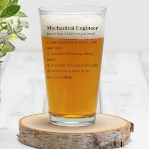 May include: A clear pint glass filled with amber liquid, resting on a wooden coaster. The glass features the text "Mechanical Engineer" with a phonetic pronunciation and a definition. The definition includes three points about the profession and a reference to "Chris".