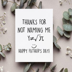 May include: A white card with black text that reads "Thanks for not naming me Y≠√π Happy Father's Day!" The card is surrounded by small white flowers and green leaves.