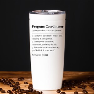 May include: A white tumbler with a black lid, featuring the text "Program Coordinator" and a definition. The definition includes phrases like "Master of calendars, chaos" and "Runs the show so smoothly." The tumbler is on a wooden surface with coffee beans.