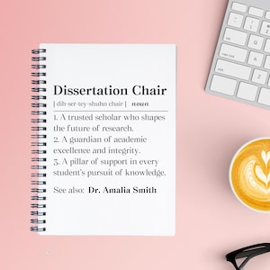 May include: A white spiral-bound notebook with the definition of "Dissertation Chair" printed on the page. The definition includes the pronunciation, part of speech, and three descriptive points. A keyboard, a cup of coffee, and glasses are also visible.