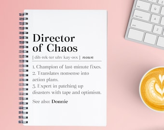 May include: A white notebook with a black spiral binding. The notebook has a black and white definition of "Director of Chaos" with the pronunciation and definition listed below. The definition includes the text "Champion of last-minute fixes", "Translates nonsense into action plans", and "Expert in patching up disasters with tape and optimism".
