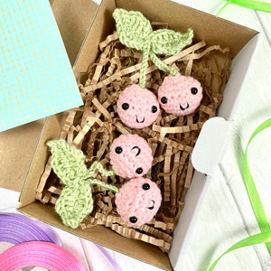 Könnte beinhalten: Ein Set aus drei gehäkelten Kirschen in einer braunen Schachtel mit zerknülltem Papier. Die Kirschen sind rosa mit schwarzen Augen und einem lächelnden Gesicht. Die Kirschen haben grüne gehäkelte Stiele und Blätter.