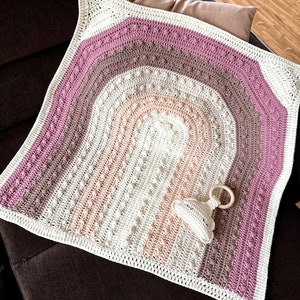 May include: A crocheted baby blanket with a rainbow design in shades of pink, brown, and white. The blanket is on a brown couch and there is a wooden teething ring and a crocheted toy next to it.