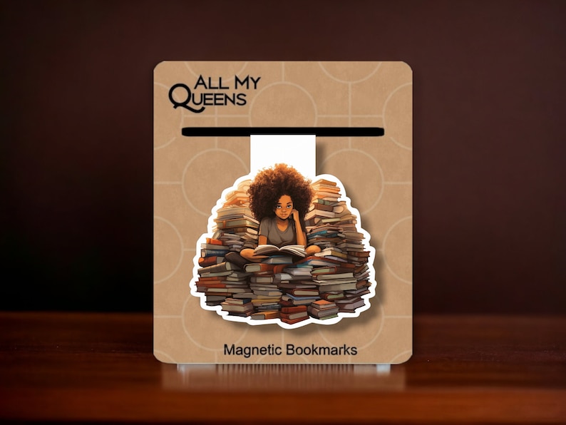 May include: A magnetic bookmark featuring a woman with afro hair sitting on a pile of books, reading a book. The bookmark is white with a brown background and the text "All My Queens" and "Magnetic Bookmarks".