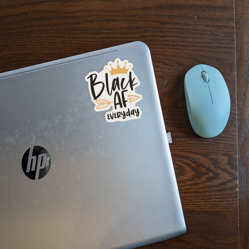 May include: A silver laptop computer with a black and gold sticker that says "Black AF everyday" with a crown and arrow. The laptop is on a wooden surface with a light blue computer mouse.