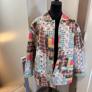 Puede incluir: Una chaqueta patchwork con cuello y cierre de botones. La chaqueta presenta una variedad de patrones de tela, incluyendo cuadros, flores y colores lisos en tonos de rosa, verde, marrón y azul. La chaqueta se muestra en un maniquí negro.