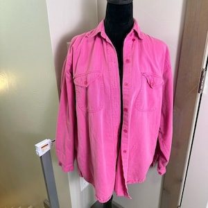 May include: A vibrant pink button-down shirt with two chest pockets. The long-sleeved shirt is displayed on a black mannequin. The shirt has a classic collar and button closures.
