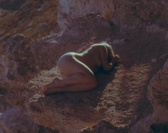 May include: A nude person lying on a rocky cliff overlooking a valley. The person is lying on their stomach with their head turned to the side. The cliff is made of light brown rock and the valley is covered in green vegetation.