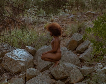 May include: A person with an afro hairstyle sits on a pile of rocks in a forest setting. The person is nude and is looking down.