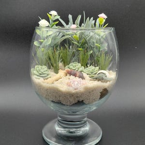 May include: A clear glass goblet terrarium filled with beige sand, featuring artificial green succulents, tall thin grasses, and small white and pink flowers. The arrangement includes a piece of light brown driftwood and a small seashell.