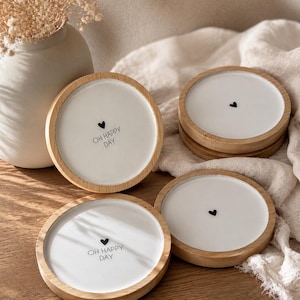Könnte beinhalten: Ein Set aus vier runden Untersetzern mit hellem Holzrand und weißer Mitte. Zwei Untersetzer zeigen ein kleines schwarzes Herzsymbol über dem Text "OH HAPPY DAY", während die anderen beiden nur das kleine schwarze Herzsymbol mittig tragen.