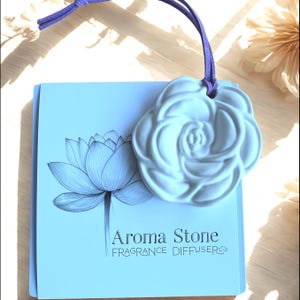 May include: A white rose-shaped aroma stone fragrance diffuser with a purple cord, resting on a light blue card. The card features a lotus flower illustration and the text "Aroma Stone FRAGRANCE DIFFUSER".
