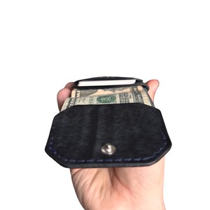Puede incluir: Una cartera de cuero negro con cierre de bot&oacute;n a presi&oacute;n y costuras azules. La cartera est&aacute; abierta, revelando un fajo de billetes estadounidenses y una tarjeta blanca insertada en el compartimento interior, mostrando su dise&ntilde;o.