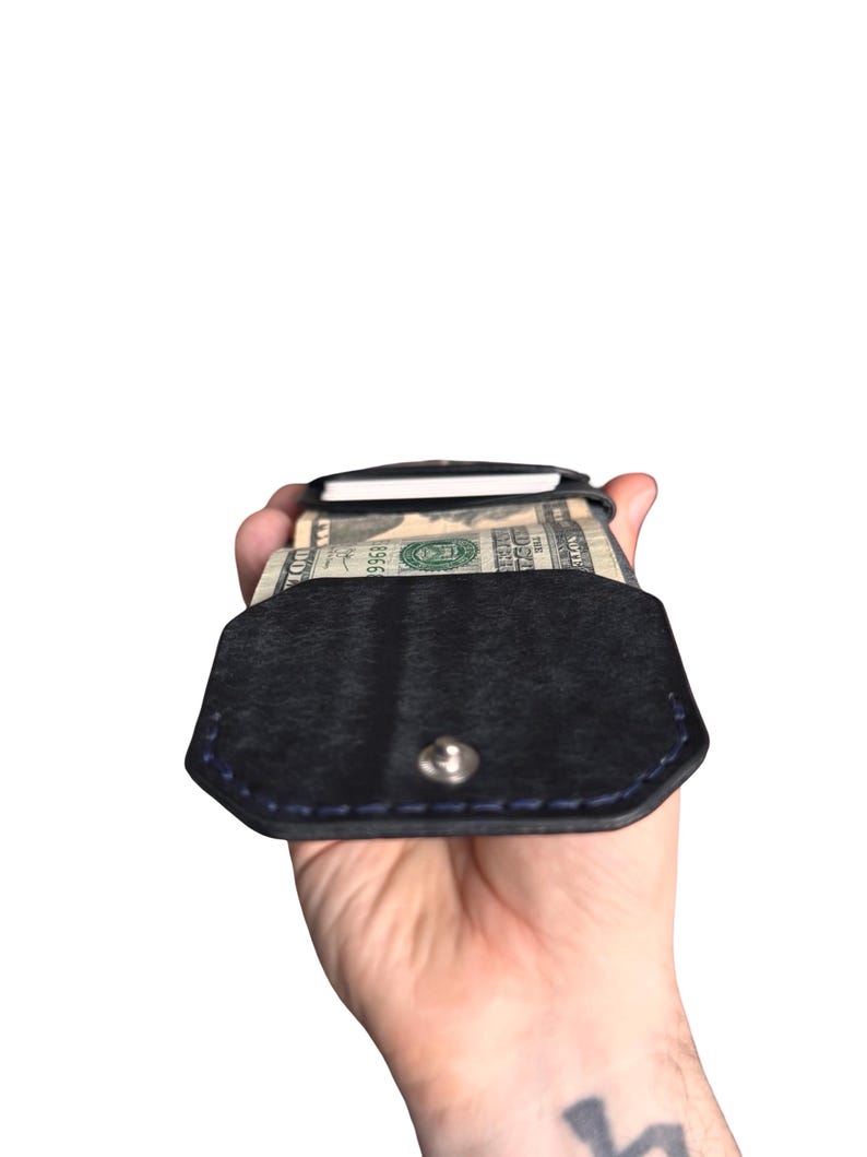 Puede incluir: Una cartera de cuero negro con cierre de bot&oacute;n a presi&oacute;n y costuras azules. La cartera est&aacute; abierta, revelando un fajo de billetes estadounidenses y una tarjeta blanca insertada en el compartimento interior, mostrando su dise&ntilde;o.