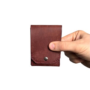 Puede incluir: Un tarjetero de cuero color burdeos oscuro con una solapa rectangular y un cierre de bot&oacute;n a presi&oacute;n de metal plateado. El accesorio presenta costuras visibles a lo largo del borde inferior y un acabado de superficie texturizado en el cuero.