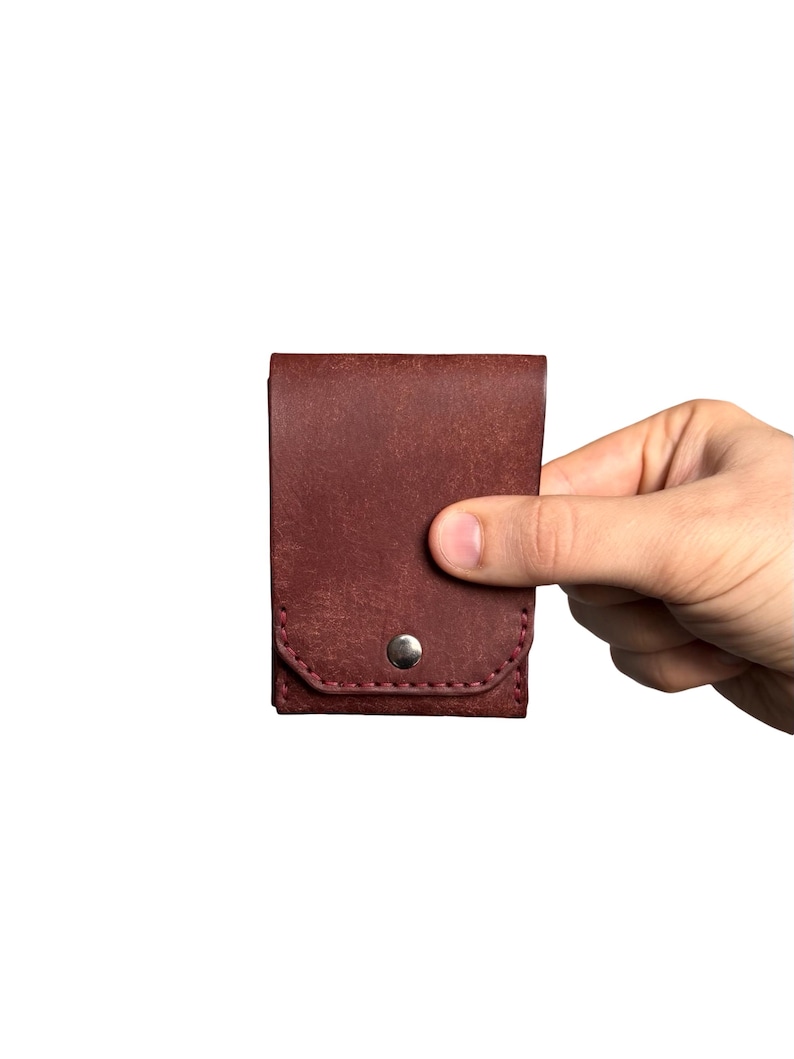 Puede incluir: Un tarjetero de cuero color burdeos oscuro con una solapa rectangular y un cierre de bot&oacute;n a presi&oacute;n de metal plateado. El accesorio presenta costuras visibles a lo largo del borde inferior y un acabado de superficie texturizado en el cuero.