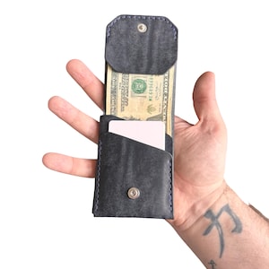 Puede incluir: Una cartera de cuero gris oscuro con costuras azules, que cuenta con un cierre de bot&oacute;n a presi&oacute;n, un bolsillo abierto con un billete de veinte d&oacute;lares y una ranura secundaria con una tarjeta blanca. La cartera tiene una orientaci&oacute;n vertical.
