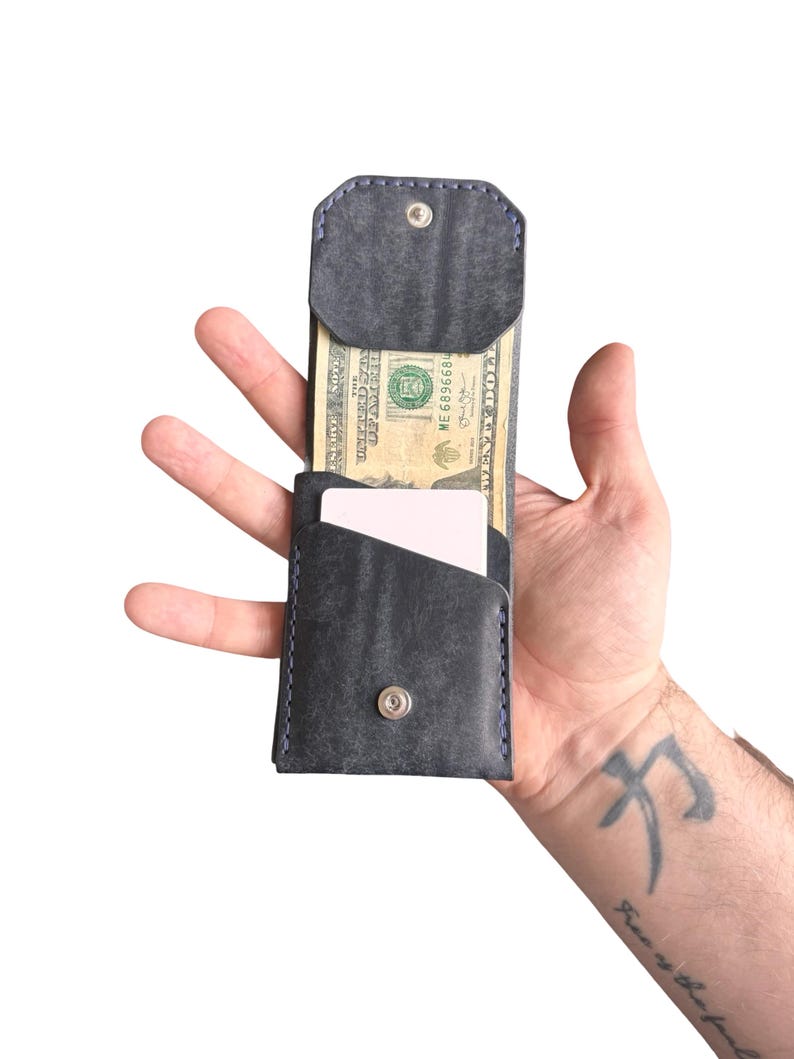 Puede incluir: Una cartera de cuero gris oscuro con costuras azules, que cuenta con un cierre de bot&oacute;n a presi&oacute;n, un bolsillo abierto con un billete de veinte d&oacute;lares y una ranura secundaria con una tarjeta blanca. La cartera tiene una orientaci&oacute;n vertical.