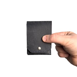 Puede incluir: Un tarjetero de cuero gris carb&oacute;n oscuro con una solapa rectangular asegurada por un bot&oacute;n de presi&oacute;n de metal plateado. El accesorio presenta costuras oscuras visibles a lo largo del borde inferior de la solapa y el cuerpo principal.