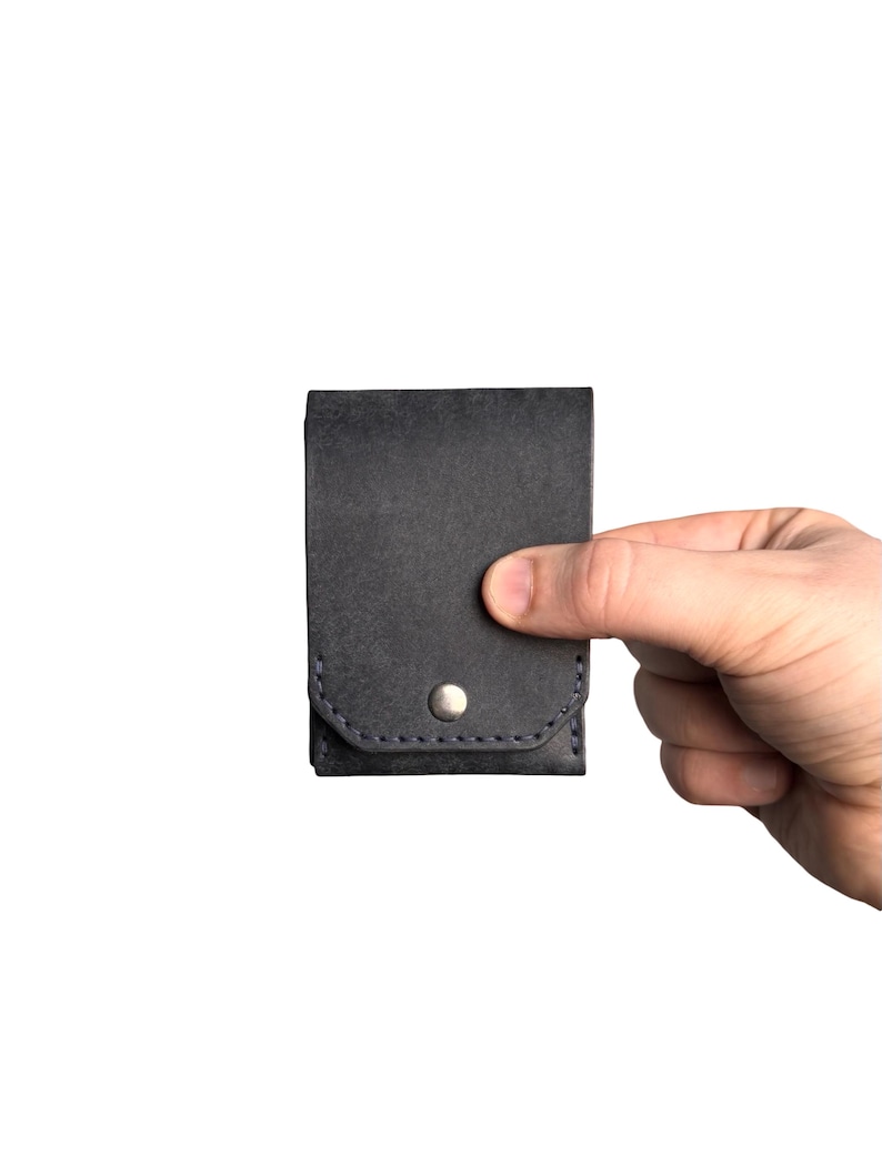 Puede incluir: Un tarjetero de cuero gris carb&oacute;n oscuro con una solapa rectangular asegurada por un bot&oacute;n de presi&oacute;n de metal plateado. El accesorio presenta costuras oscuras visibles a lo largo del borde inferior de la solapa y el cuerpo principal.