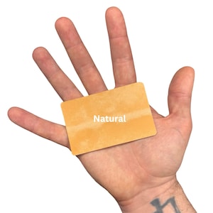 Puede incluir: Una tarjeta rectangular con una superficie texturizada de color naranja claro que presenta la palabra Natural impresa en blanco en el centro. La tarjeta tiene esquinas redondeadas y un acabado mate, con un tono uniforme en toda su superficie.