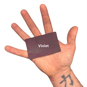 Puede incluir: Una peque&ntilde;a tarjeta rectangular de color violeta oscuro con esquinas redondeadas. La palabra Violet est&aacute; impresa en letras may&uacute;sculas blancas sin remates en el centro de la tarjeta. La tarjeta se sostiene plana contra una palma abierta.