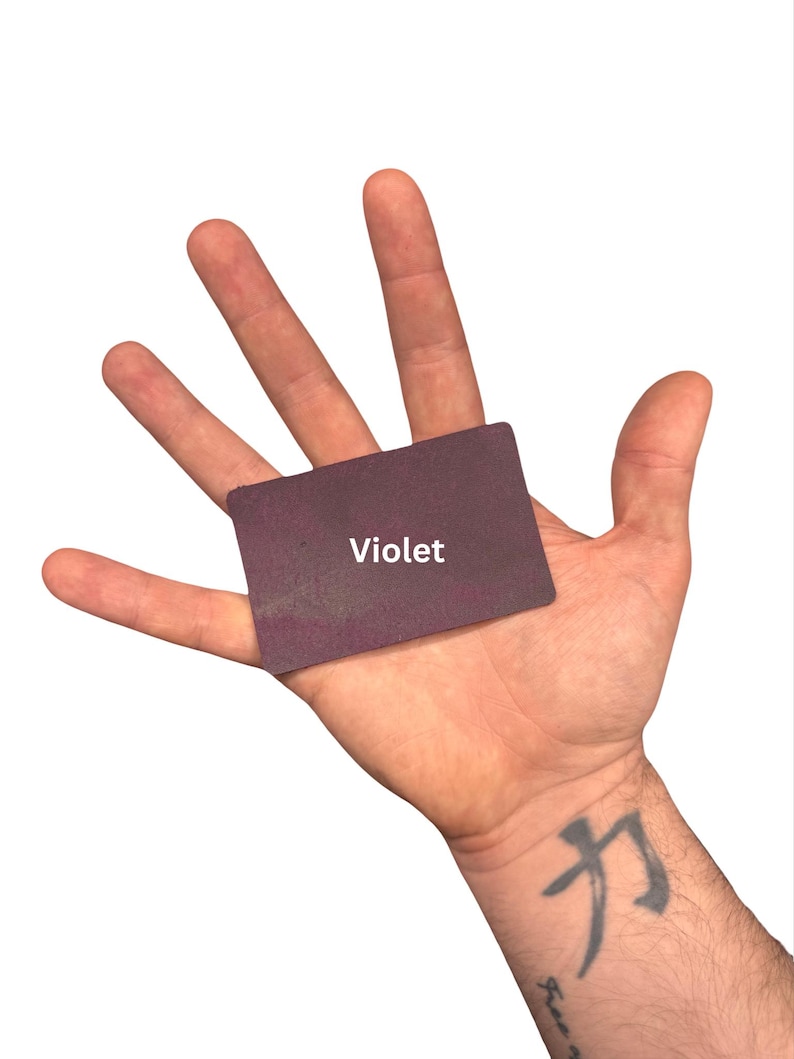 Puede incluir: Una peque&ntilde;a tarjeta rectangular de color violeta oscuro con esquinas redondeadas. La palabra Violet est&aacute; impresa en letras may&uacute;sculas blancas sin remates en el centro de la tarjeta. La tarjeta se sostiene plana contra una palma abierta.