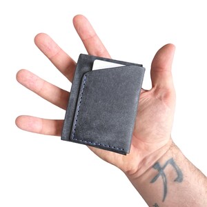 Puede incluir: Un tarjetero de color gris carb&oacute;n con forma rectangular y costuras azules visibles a lo largo del borde lateral. El tarjetero tiene una abertura superior inclinada que revela una tarjeta blanca insertada en el compartimento principal.