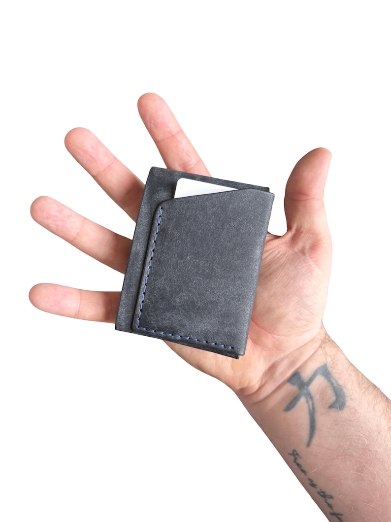 Puede incluir: Un tarjetero de color gris carb&oacute;n con forma rectangular y costuras azules visibles a lo largo del borde lateral. El tarjetero tiene una abertura superior inclinada que revela una tarjeta blanca insertada en el compartimento principal.