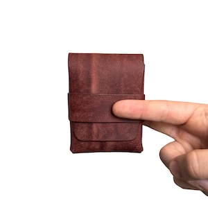 May include: A rectangular, dark reddish-brown leather card holder featuring a smooth, matte finish and a horizontal strap across the center. The item has a simple, folded construction with visible grain texture and clean, straight edges throughout.