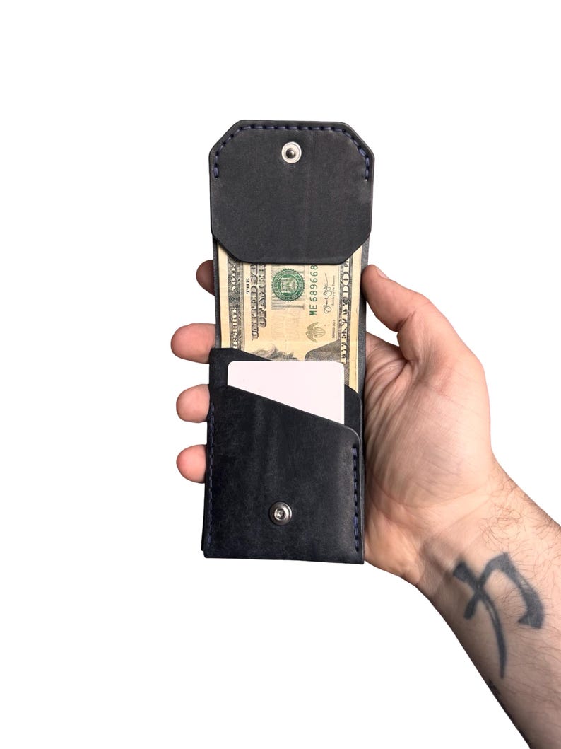 Puede incluir: Una cartera de cuero azul marino oscuro con una solapa plegable con cierre de bot&oacute;n a presi&oacute;n de metal plateado, un bolsillo vertical que contiene un billete de veinte d&oacute;lares y una ranura frontal con una tarjeta blanca.
