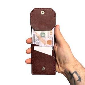 Puede incluir: Una cartera de cuero marr&oacute;n oscuro con cierre de solapa asegurado por un bot&oacute;n de presi&oacute;n de metal plateado. La cartera contiene un billete de diez d&oacute;lares y una tarjeta rectangular blanca. Presenta costuras visibles en los bordes.