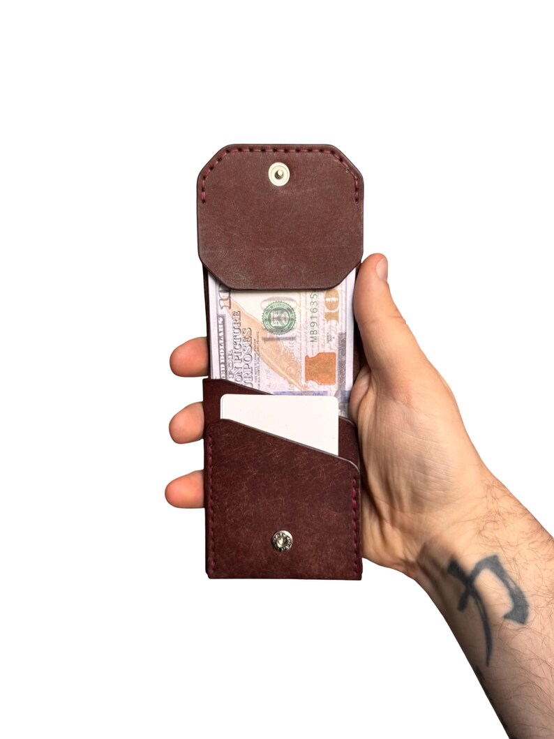 Puede incluir: Una cartera de cuero marr&oacute;n oscuro con cierre de solapa asegurado por un bot&oacute;n de presi&oacute;n de metal plateado. La cartera contiene un billete de diez d&oacute;lares y una tarjeta rectangular blanca. Presenta costuras visibles en los bordes.