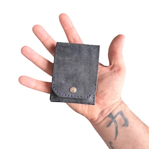 Puede incluir: Una cartera peque&ntilde;a y rectangular de color gris oscuro con una superficie texturizada. La cartera cuenta con un cierre de solapa asegurado por un bot&oacute;n de presi&oacute;n de metal plateado. Los bordes est&aacute;n cosidos con hilo azul oscuro.