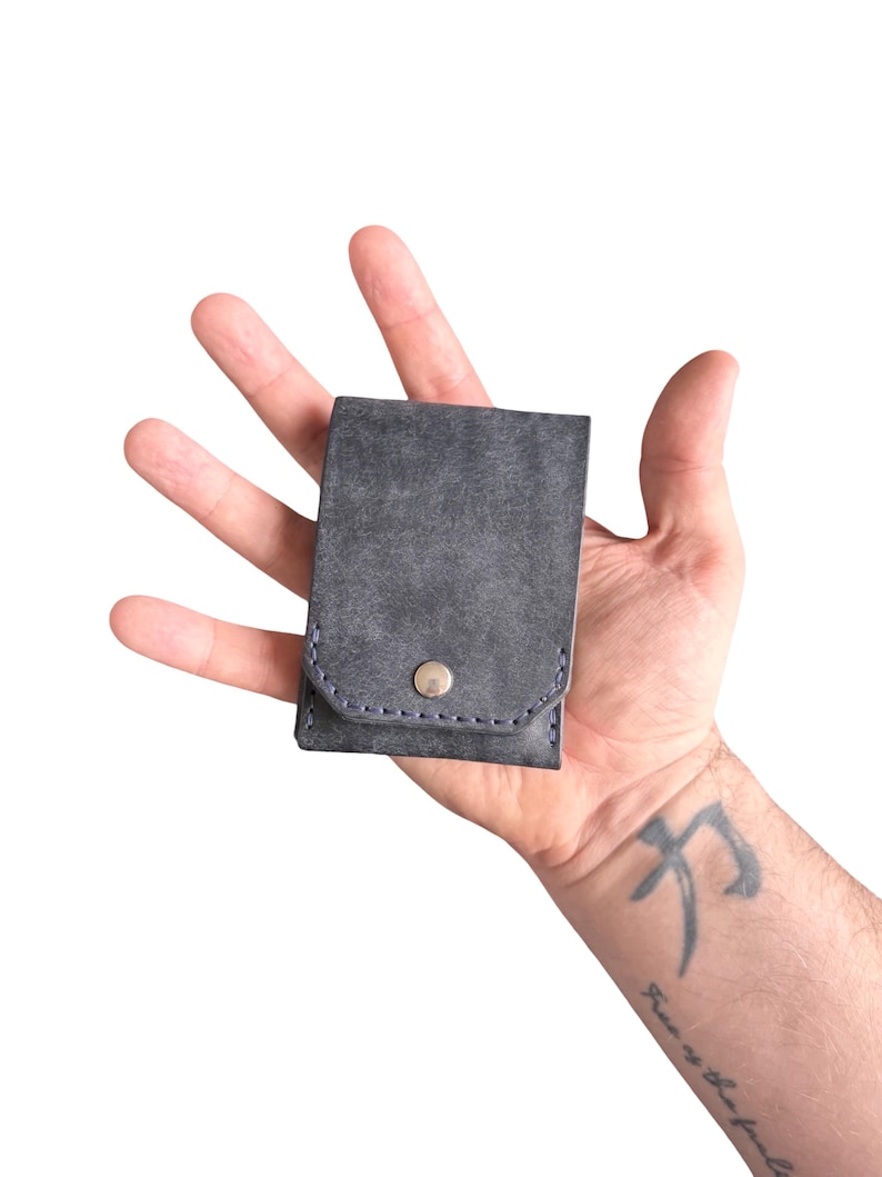 Puede incluir: Una cartera peque&ntilde;a y rectangular de color gris oscuro con una superficie texturizada. La cartera cuenta con un cierre de solapa asegurado por un bot&oacute;n de presi&oacute;n de metal plateado. Los bordes est&aacute;n cosidos con hilo azul oscuro.