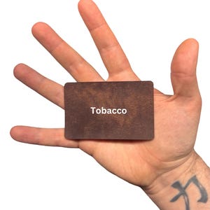 Puede incluir: Una tarjeta rectangular con una superficie texturizada de color marr&oacute;n oscuro que se asemeja al cuero. La palabra Tobacco est&aacute; impresa en letras may&uacute;sculas blancas sin remates en el centro de la tarjeta. Tiene esquinas redondeadas y un perfil plano.
