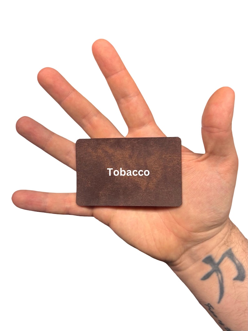 Puede incluir: Una tarjeta rectangular con una superficie texturizada de color marr&oacute;n oscuro que se asemeja al cuero. La palabra Tobacco est&aacute; impresa en letras may&uacute;sculas blancas sin remates en el centro de la tarjeta. Tiene esquinas redondeadas y un perfil plano.