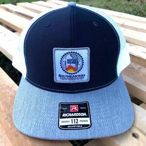 May include: A black and white trucker hat with a gray brim. The hat has a patch on the front that says "Southeastern Pipe Fabrication, Petite Anse, LA". The hat is made by Richardson and is adjustable.