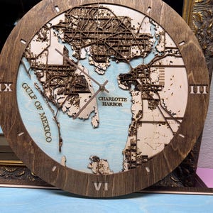 May include: Wooden wall clock depicting a detailed map of Charlotte Harbor, Florida, with Roman numerals. The clock has a light blue background representing the water, and the text "Gulf of Mexico" is visible.