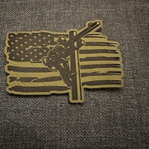 May include: A green and brown patch with a silhouette of a person climbing a utility pole in front of an American flag.