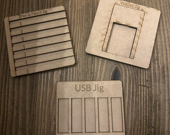 May include: Three wooden jigs for crafting. The first jig is labeled "Pen & Pencil Jig" and has multiple slots for holding pens and pencils. The second jig is labeled "Wallet Jig" and has a rectangular cutout for a wallet. The third jig is labeled "USB Jig" and has five slots for holding USB drives.