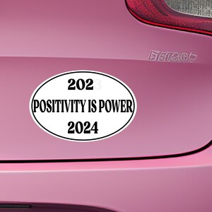 May include: White oval sticker with black text that reads "2023 POSITIVITY IS POWER 2024" on a pink car.