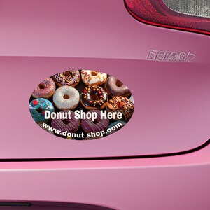 May include: A pink car with a donut shop sticker on the back. The sticker is oval shaped and features a variety of donuts with colorful frosting and sprinkles. The text on the sticker reads "Donut Shop Here" and "www.donutshop.com".
