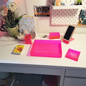 Peut inclure: Un bureau blanc avec des organiseurs en acrylique rose, un plateau rose et un téléphone portable noir sur un support. Le bureau a un tiroir et une étagère en dessous. Il y a un carnet avec une citrouille sur la couverture et un bloc-notes avec le mot "2023" sur la couverture.