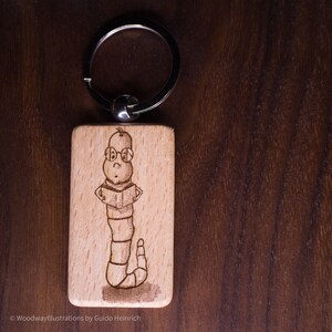 Puede incluir: Llavero de madera con una ilustración grabada con láser de un gusano con gafas leyendo un libro. El llavero está unido a un anillo de metal plateado.