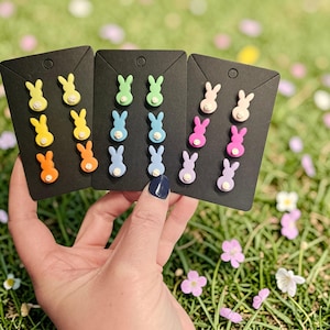 May include: A hand holds three black cards displaying colorful bunny-shaped stud earrings. The earrings are in various colors, including yellow, orange, green, blue, pink, and purple, with white accents. The background is a blurred green field with flowers.