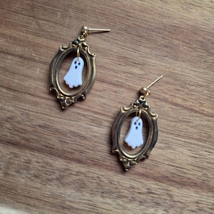 May include: A pair of dangle earrings featuring small white ghost charms suspended within ornate, antique-style oval gold-toned metal frames. Each earring is attached to a gold-toned post backing, showcasing a detailed, vintage-inspired design.