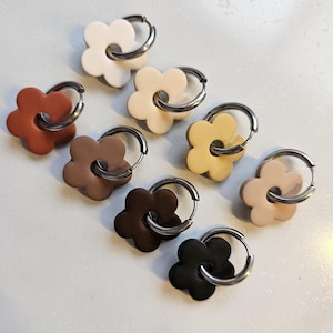 May include: A selection of flower-shaped earrings in shades of white, beige, brown, and black. Each earring has a silver-coloured hoop. The earrings are displayed on a light-coloured surface.