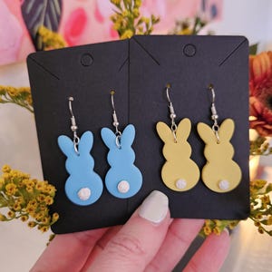 Puede incluir: Un par de pendientes azules y un par amarillos con forma de conejito. Cada pendiente tiene una cola redonda blanca y ganchos plateados. Los pendientes se muestran en tarjetas negras.